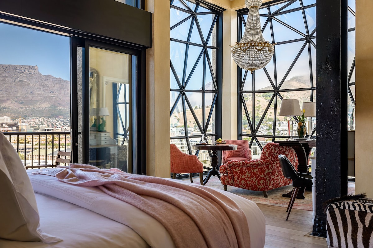 A bedroom with a large bed, a view of a cityscape and mountains through large windows, a chandelier, and a balcony with chairs and a table.