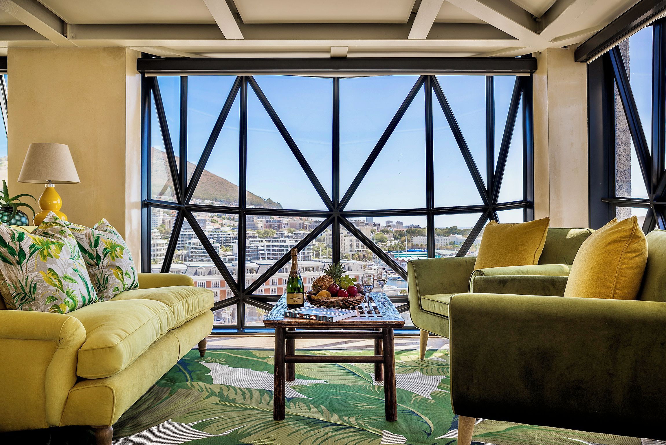 Una habitación con un sofá amarillo, un sillón verde y una mesa de centro con una botella y fruta.