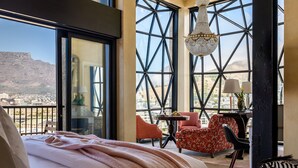A bedroom with a large bed, a view of a cityscape and mountains through large windows, a chandelier, and a balcony with chairs and a table. - The Silo Hotel (Cape Town)