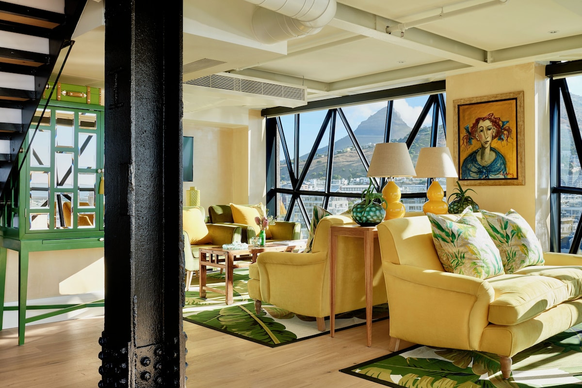 A modern living room with a yellow sofa, a green accent wall, and a large window offering a view of a cityscape and mountains.