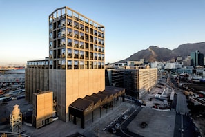 A modern building with a unique geometric facade, surrounded by a busy urban area with a mountain in the background. - The Silo Hotel (Cape Town)