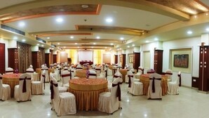 Un salón de banquetes con varias mesas redondas, cada una rodeada de sillas con cintas. El salón tiene un techo alto con iluminación empotrada y una zona de escenario central.