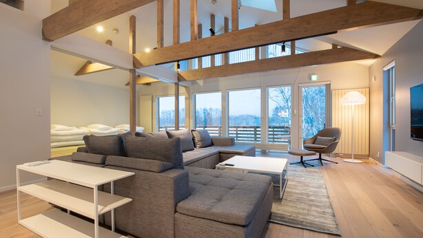 Ein modernes Wohnzimmer mit Ecksofa, weißem Couchtisch und Stehlampe. Ein großes Fenster bietet Blick ins Freie.