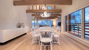 Ein moderner Essbereich mit einem runden Tisch und weißen Stühlen, Holzbalken an der Decke und großen Fenstern mit Blick auf Bäume.