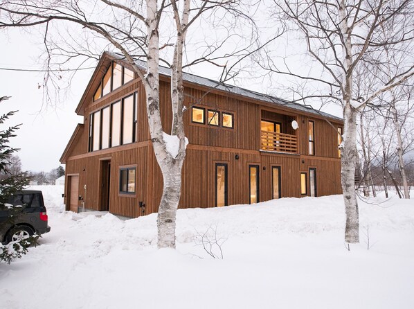 A large, multi-story wooden house with a balcony, surrounded by snow-covered ground and bare trees. - Yoteiyama House (Kutchan)