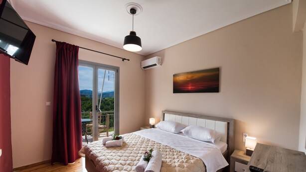 A hotel room with a bed, a TV, an air conditioner, a painting, and a balcony with red curtains. - Magicians Apartments (Lefkada)