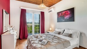 A bedroom with a bed, a dresser, a window with curtains, a painting, and a view of the outdoors. - Magicians Apartments (Lefkada)