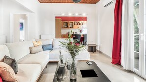 A modern living room with a white sofa, a coffee table, and a vase with flowers. There is a kitchen in the background with wooden cabinets and a red countertop. - Magicians Apartments (Lefkada)