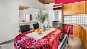 A dining area with a table set for two, a red tablecloth, and a fruit bowl. There is a wooden ceiling and a kitchen area with wooden cabinets and a red wall. - Magicians Apartments (Lefkada)