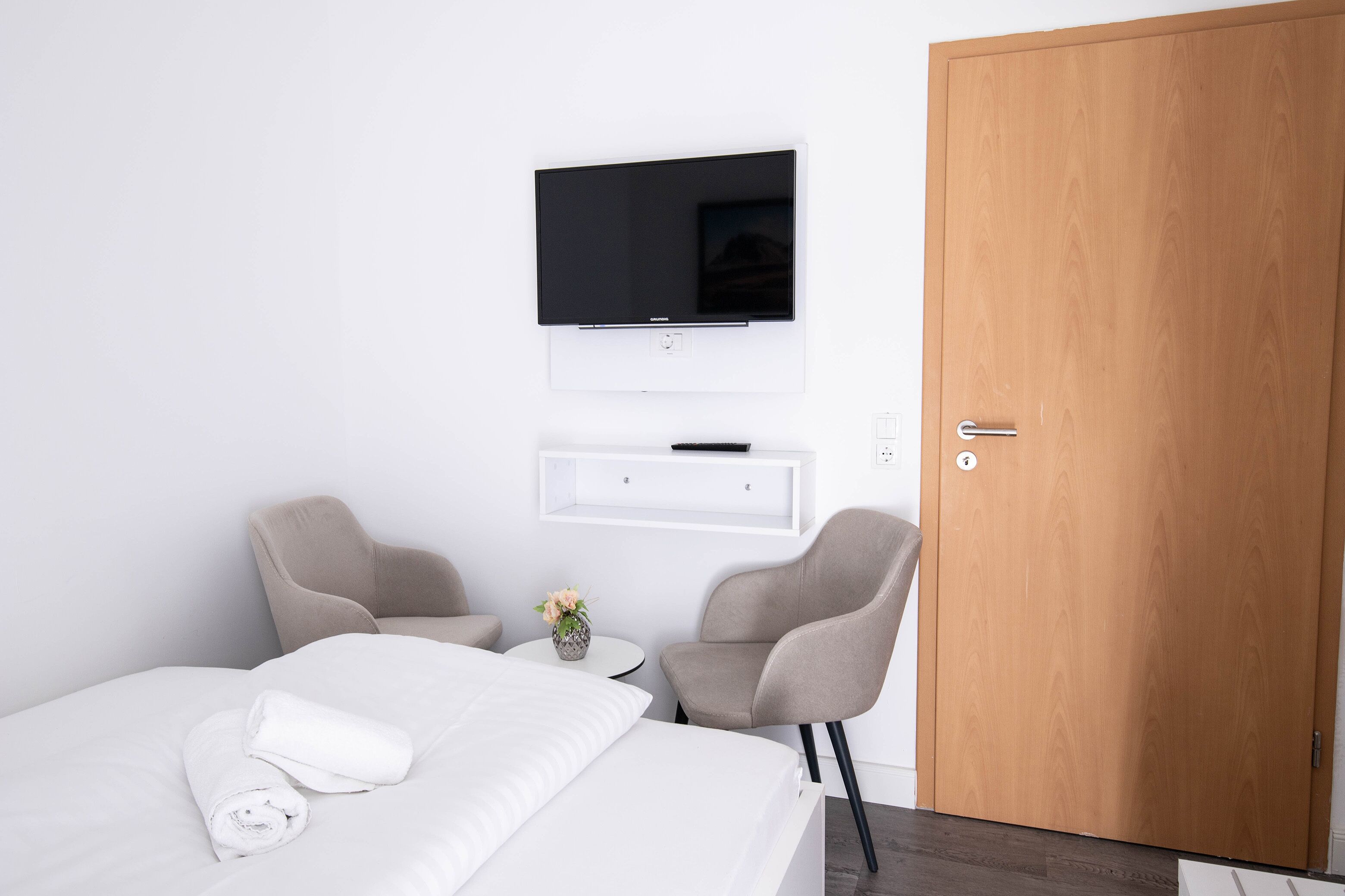 A hotel room with a bed, two chairs, a small table with a vase, a TV mounted on the wall, and a wooden door.