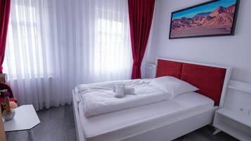 A neatly made bed with white bedding and a red headboard, a white bedside table with a vase of flowers, and a painting of a mountainous landscape on the wall.