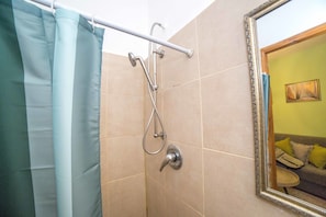 Casa de banho com chuveiro, porta-toalhas e espelho refletindo um quarto com sofá e pintura.