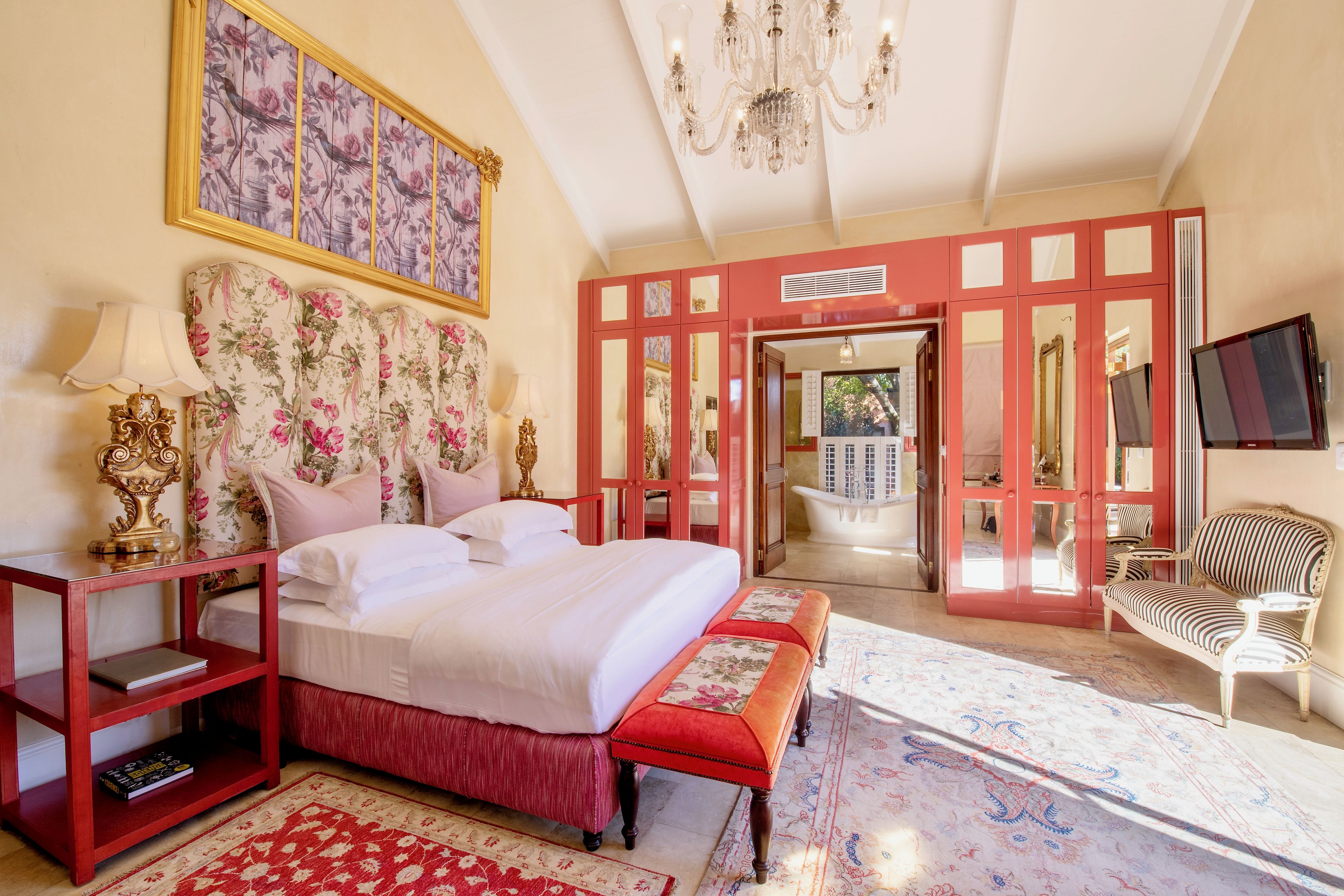 A spacious bedroom with a large bed, a sitting area with a chair, a red bench, and a view of the outside.