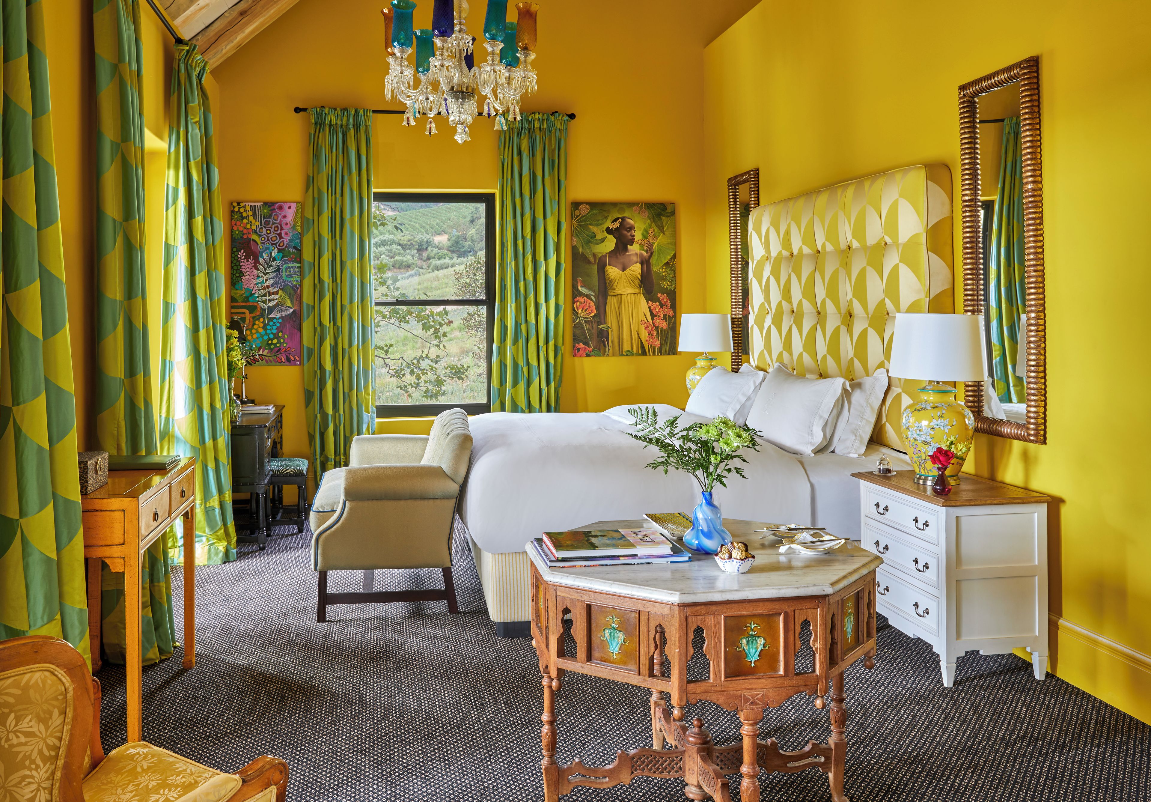 A hotel room with a bed, a desk, a chair, a small table with a vase, and a window with curtains.