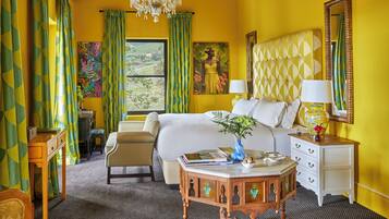 A hotel room with a bed, a desk, a chair, a small table with a vase, and a window with curtains.