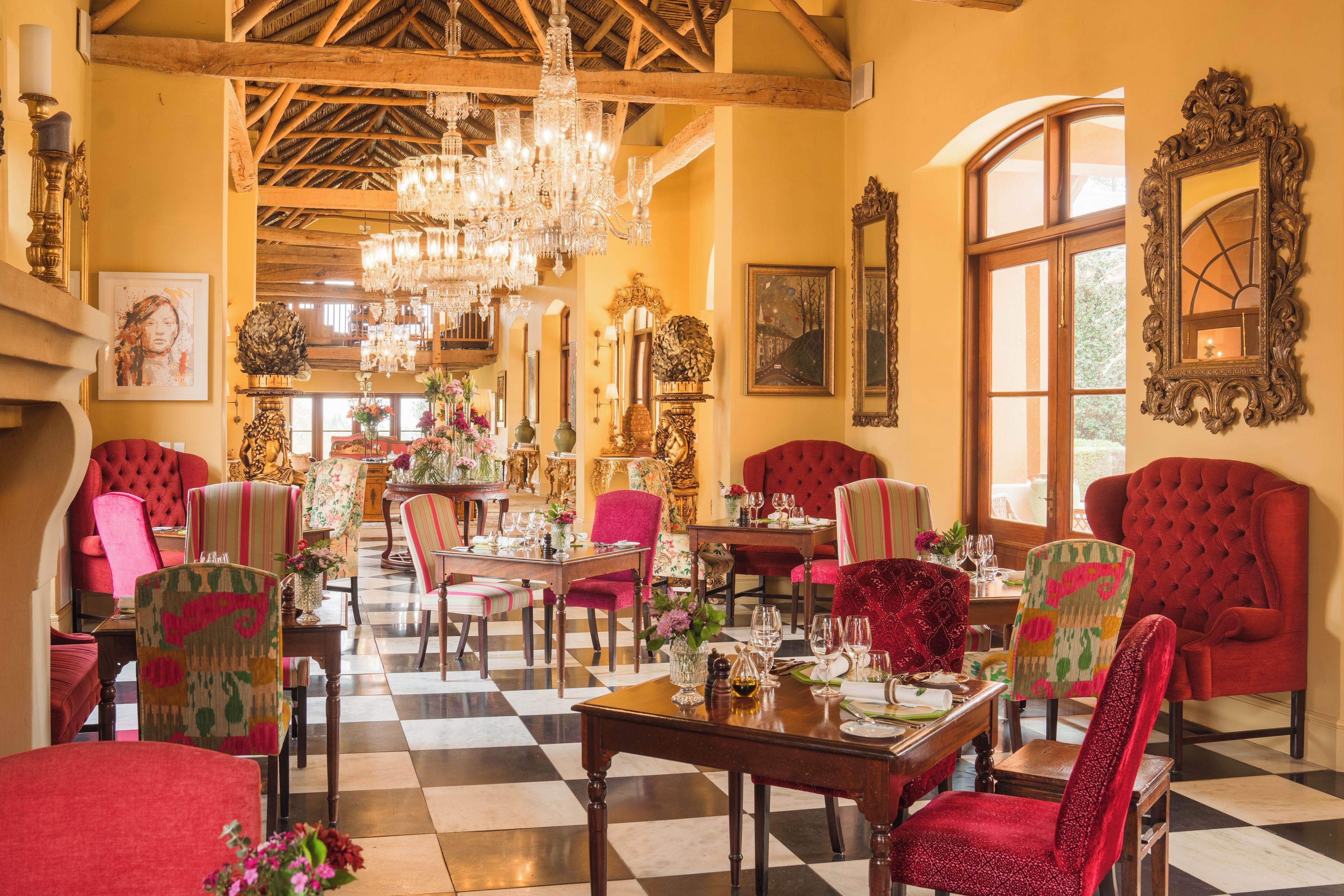 A hotel lobby with a checkered floor, red upholstered chairs, a chandelier, and a dining area set for a meal.