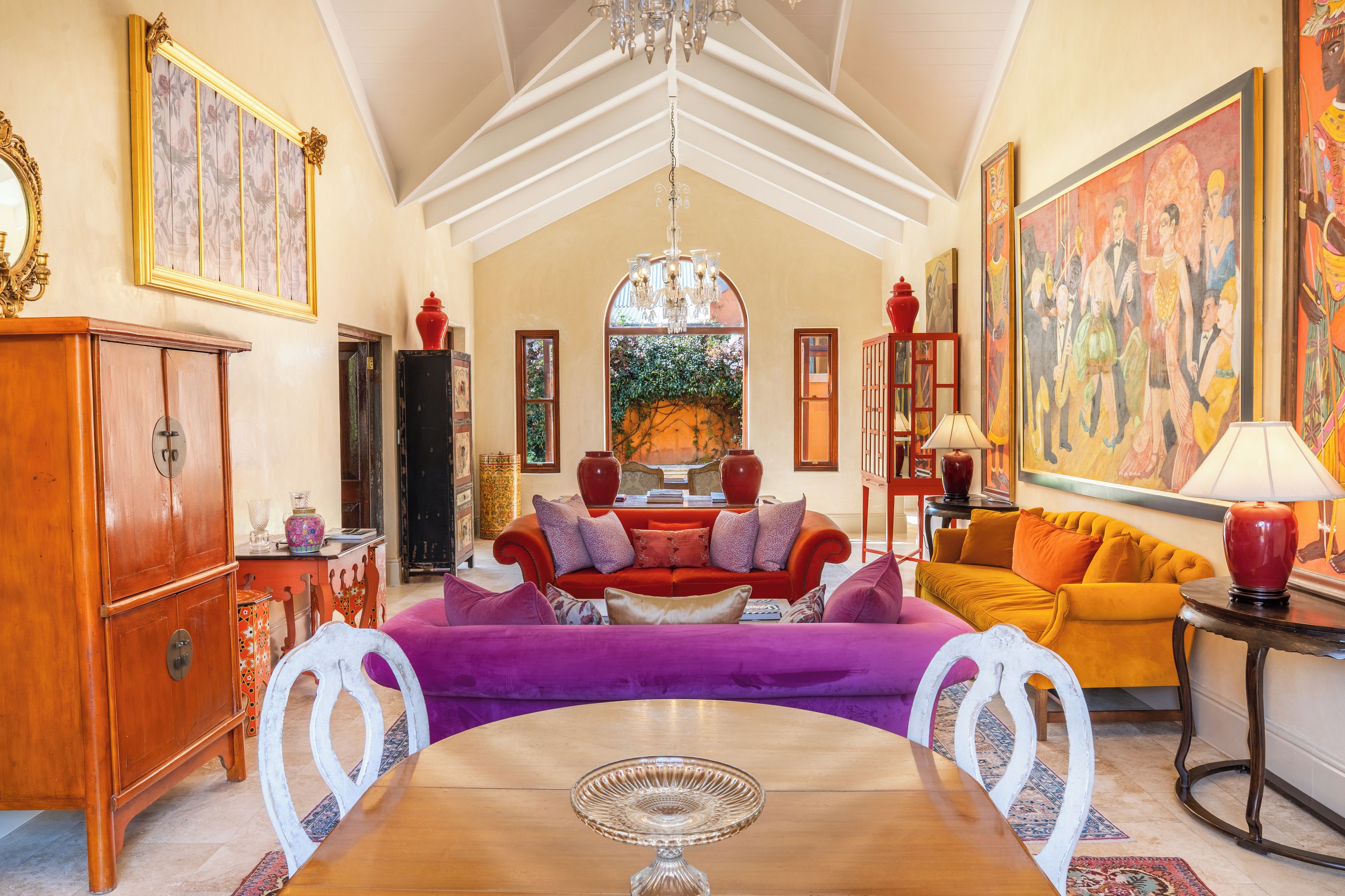 Een ruime woonkamer met een hoge plafond, een kroonluchter, een groot schilderij en een eethoek met een tafel en stoelen.