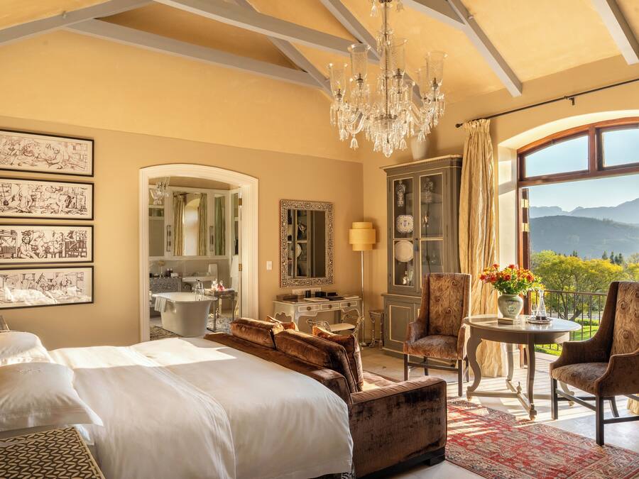 A spacious bedroom with a large bed, a sitting area, a chandelier, and a view of the outdoors.