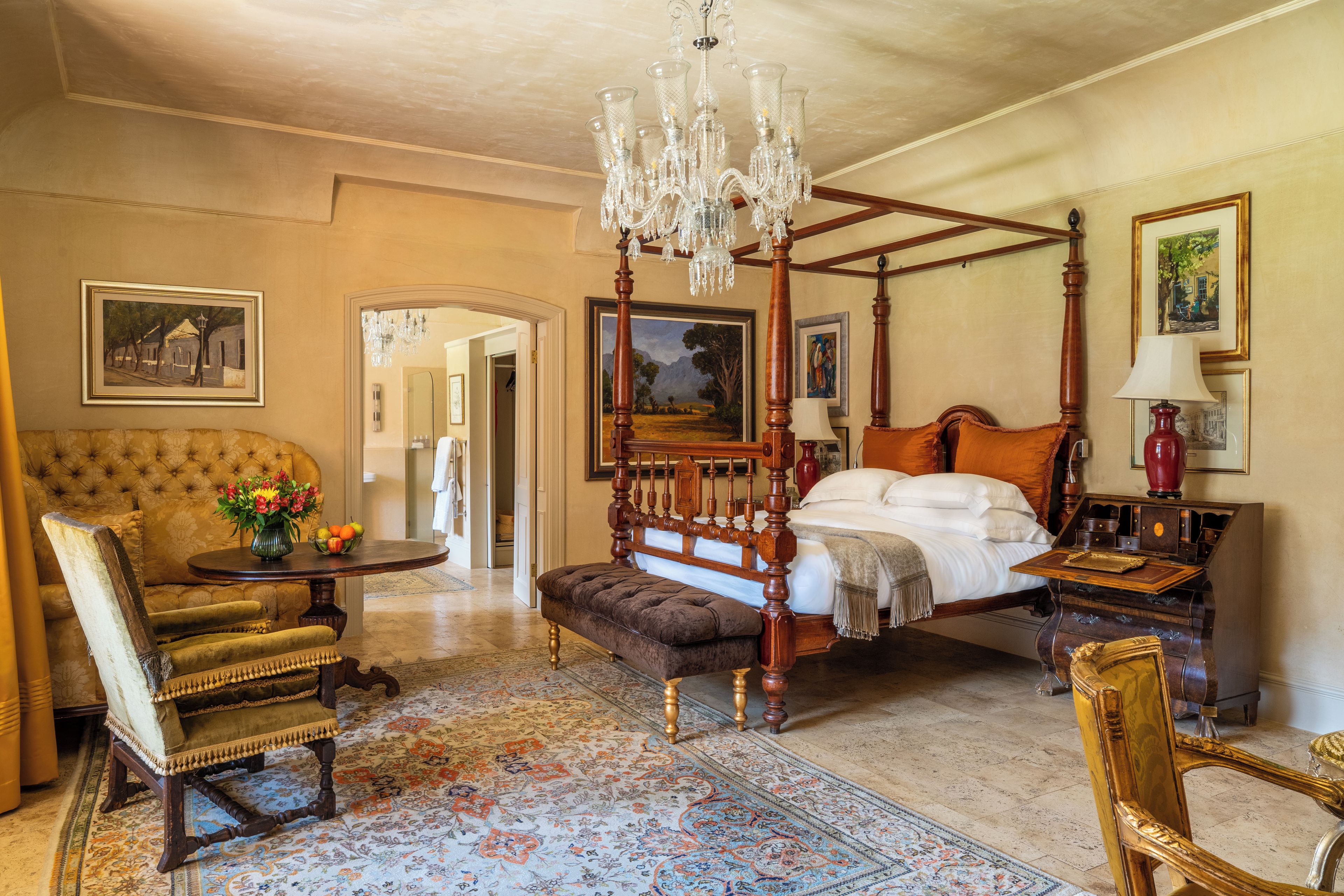 A traditional bedroom with a four-poster bed, a sitting area with a chair and table, and a chandelier.