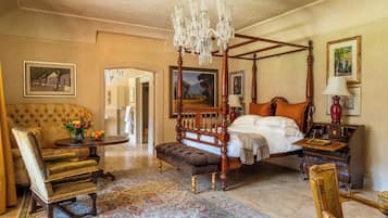 A traditional bedroom with a four-poster bed, a sitting area with a chair and table, and a chandelier.