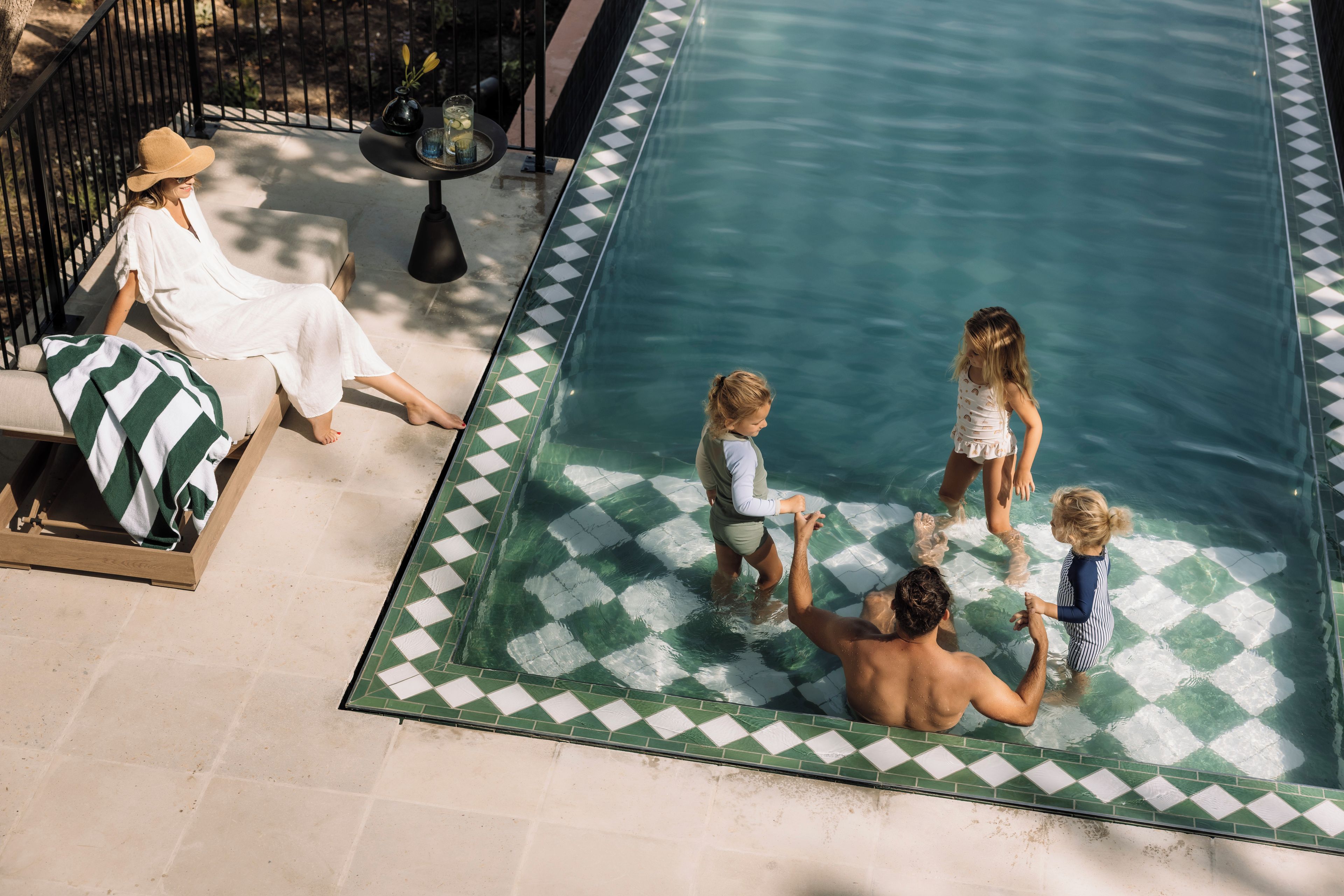 A swimming pool with a checkered edge, a lounge area with a sofa and a table, and a person sitting by the pool.