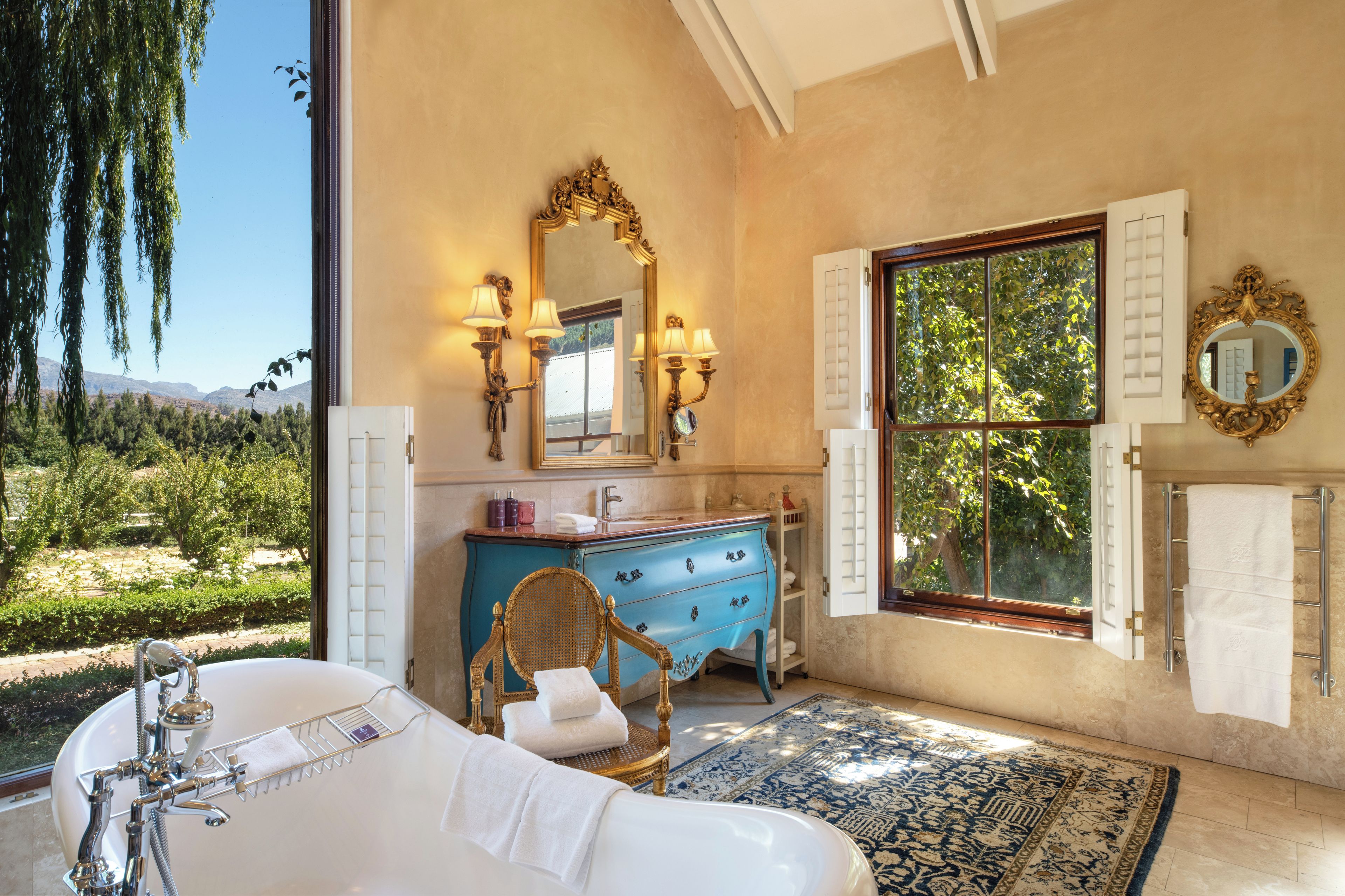 A bathroom with a bathtub, a vanity with a blue cabinet, a large mirror, and a view of greenery outside.