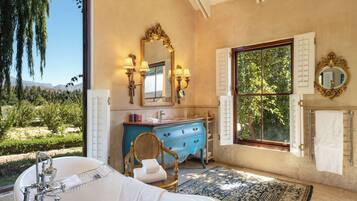 A bathroom with a bathtub, a vanity with a blue cabinet, a large mirror, and a view of greenery outside.
