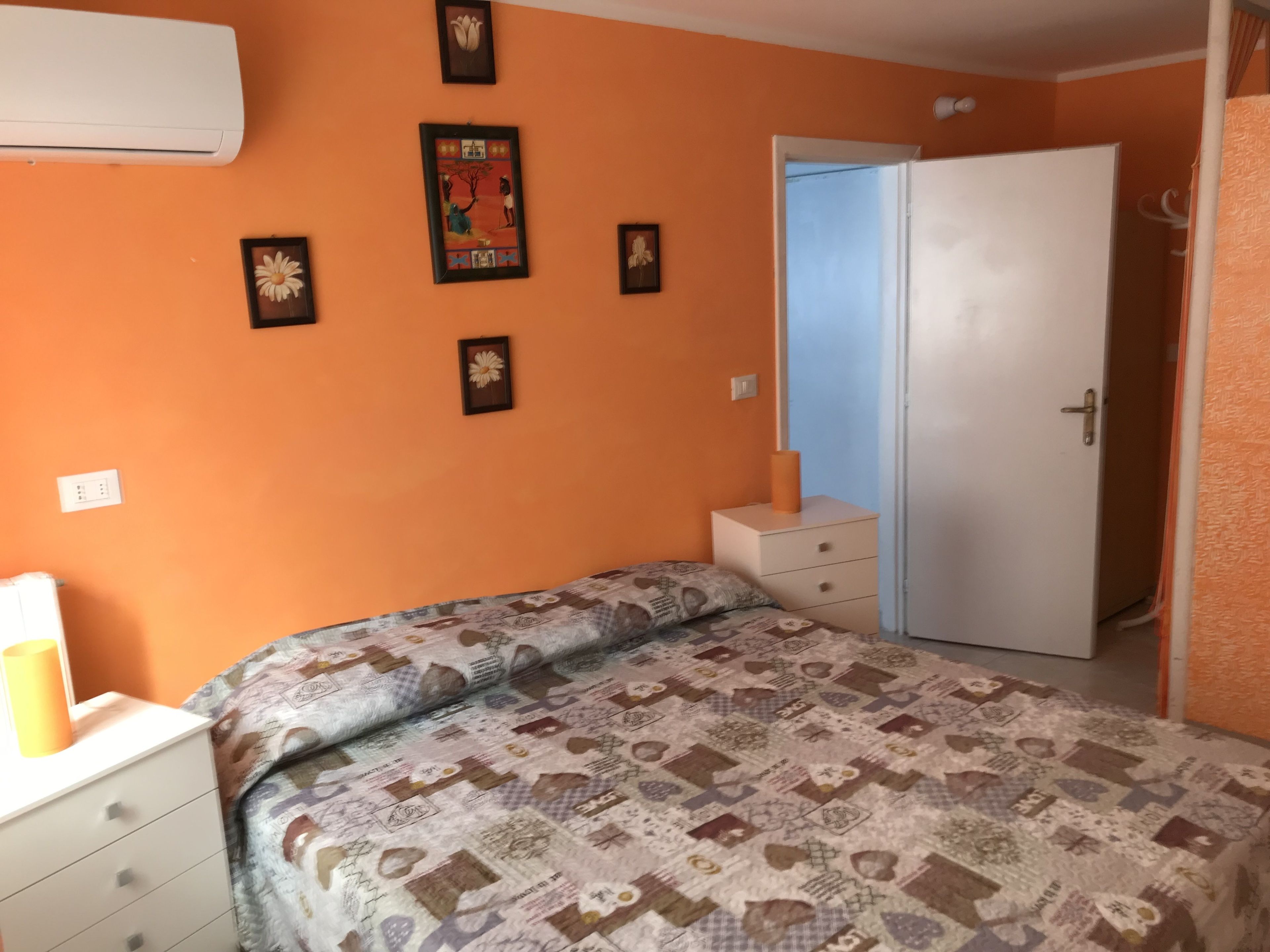 Una camera da letto con una parete arancione, un letto con copripiumino fantasia, un comodino bianco e una porta che dà su un'altra stanza.