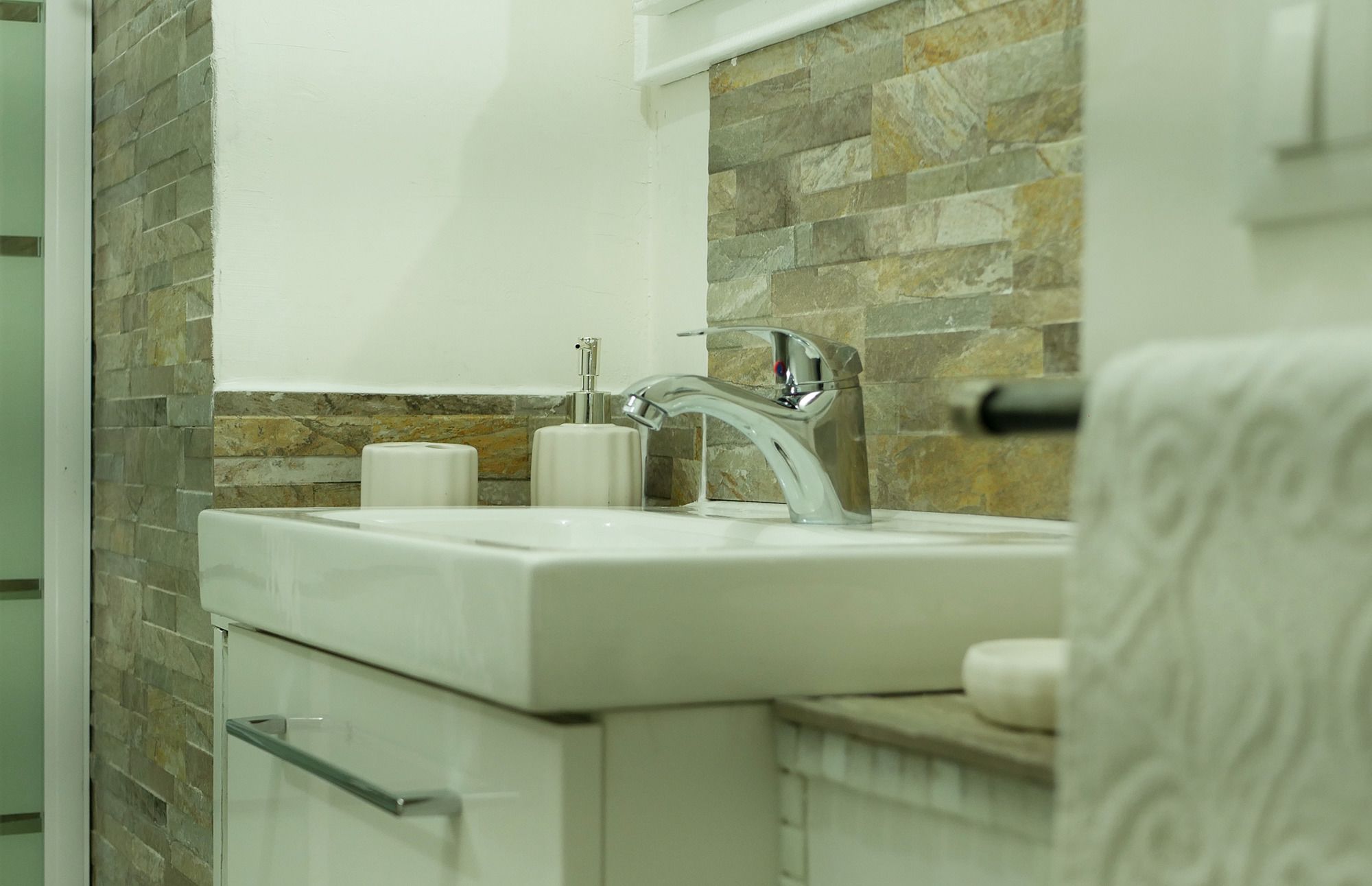 A bathroom with a white sink, a chrome faucet, and a stone backsplash. There are two white soap dispensers on the sink.