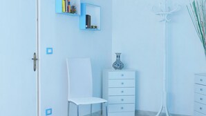 Una habitación con una silla blanca, un aparador blanco, una estantería azul con libros, un jarrón y una lámpara de pie.