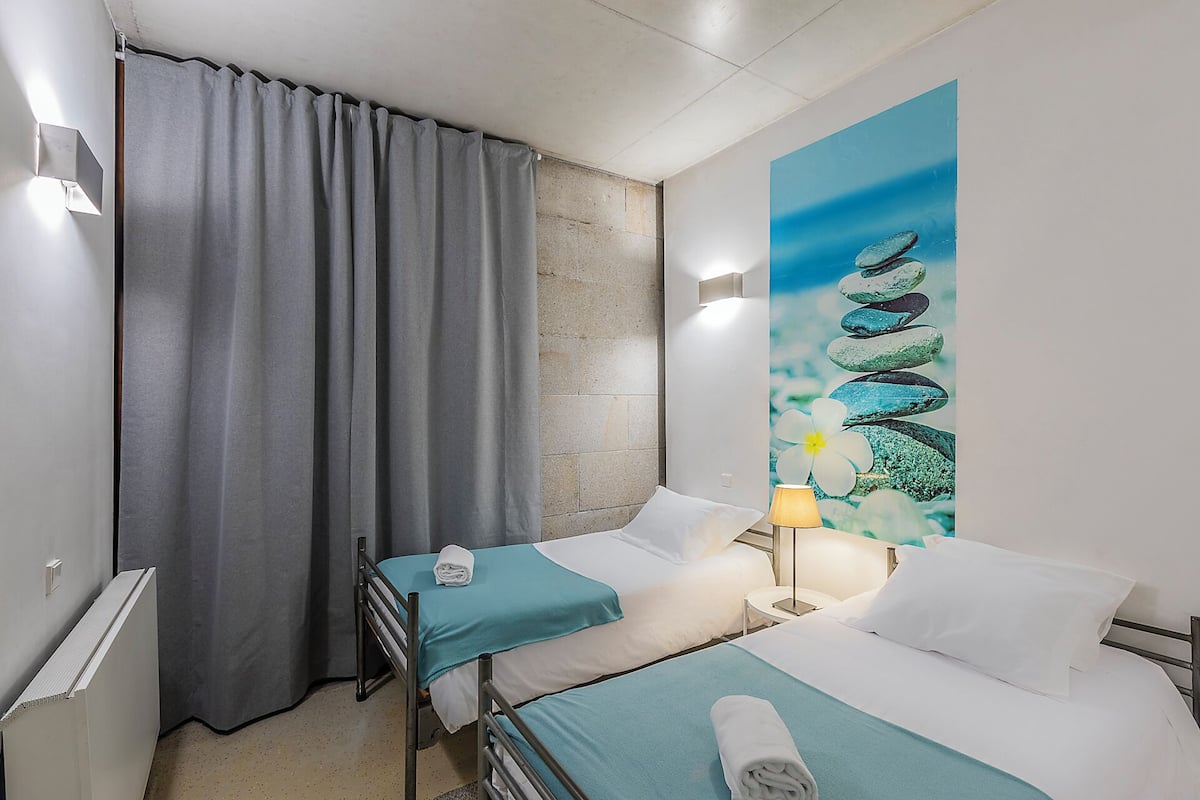 Ein Hotelzimmer mit zwei Betten, einem grauen Vorhang, einem Gemälde mit Steinen und Blumen sowie einer Lampe.