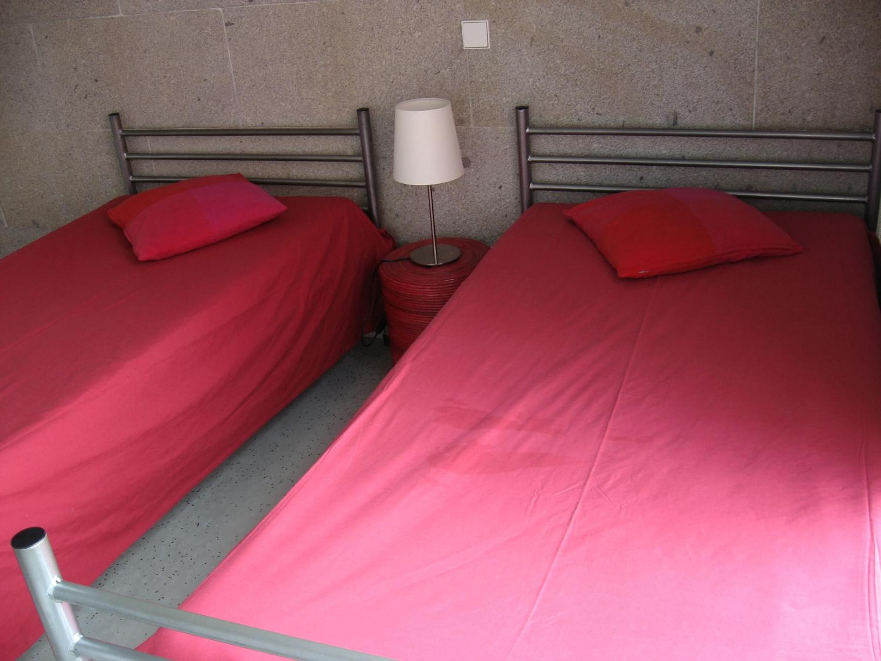 Quarto com duas camas de solteiro, cada uma com colcha e almofadas vermelhas. Uma mesa de cabeceira individual com abajur fica entre as camas.