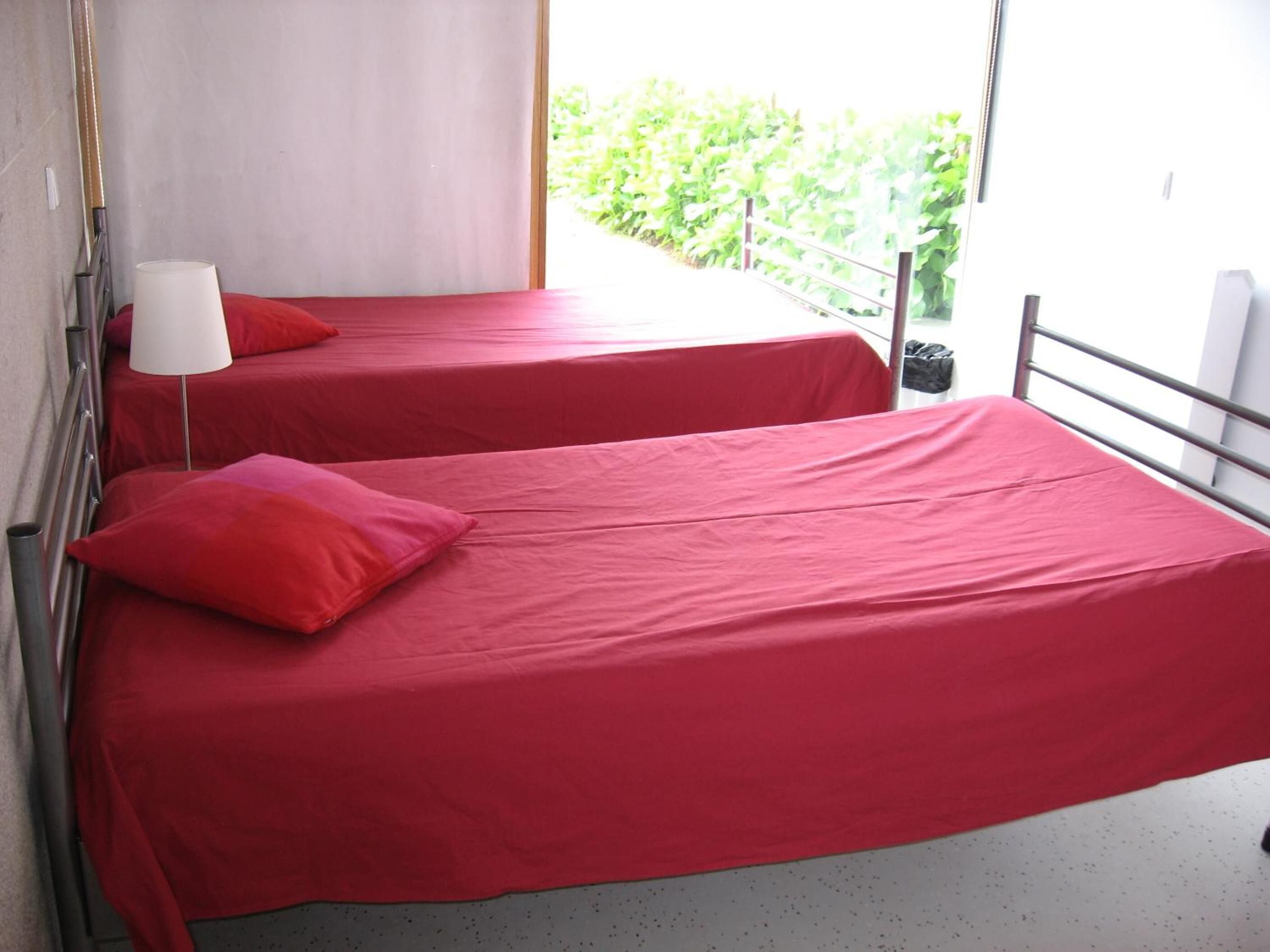 Deux lits simples avec une literie rouge, une lampe blanche et un balcon doté d’une rambarde en métal.