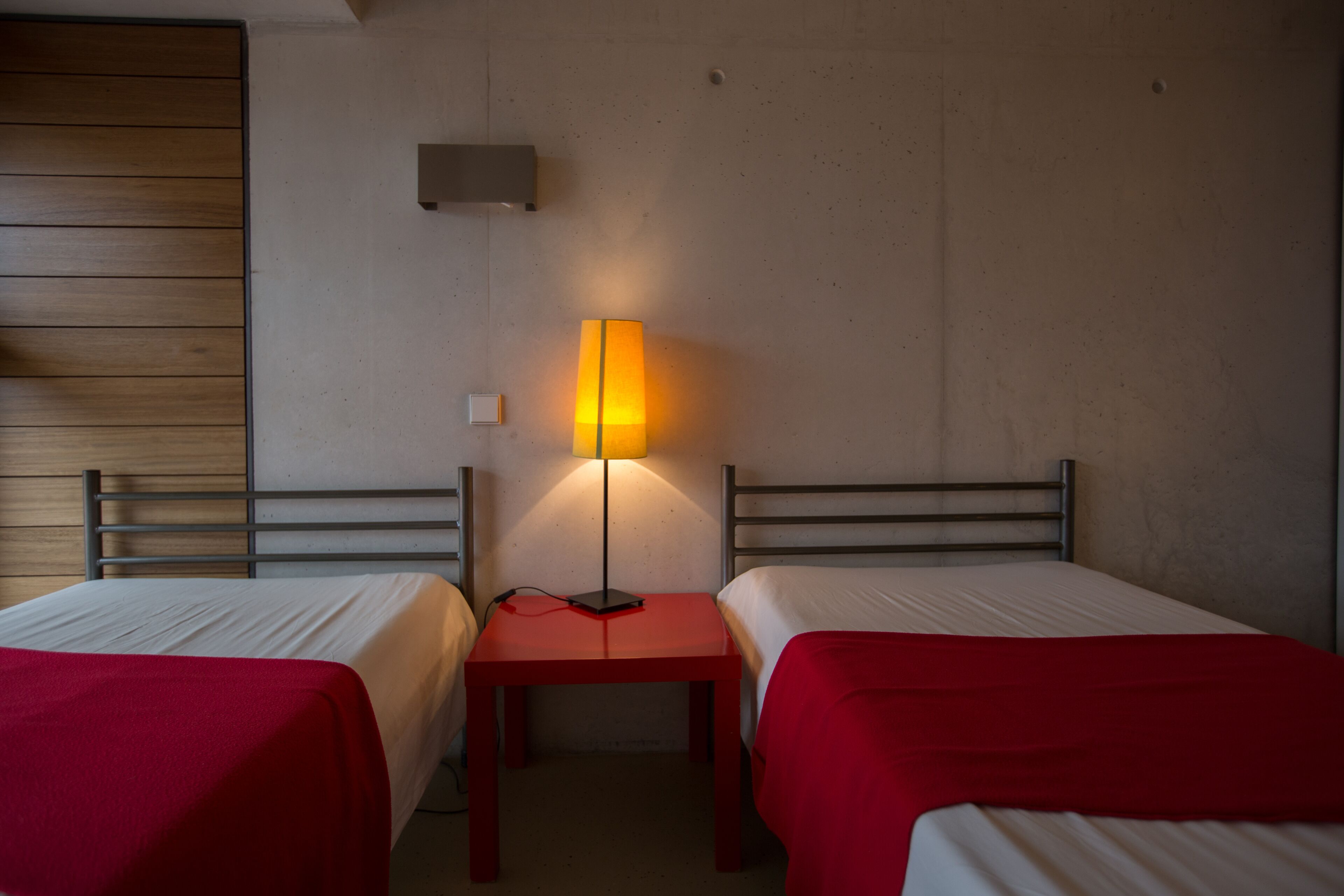 Quarto de hotel com duas camas, uma mesa vermelha, uma luminária e um painel de madeira na parede.