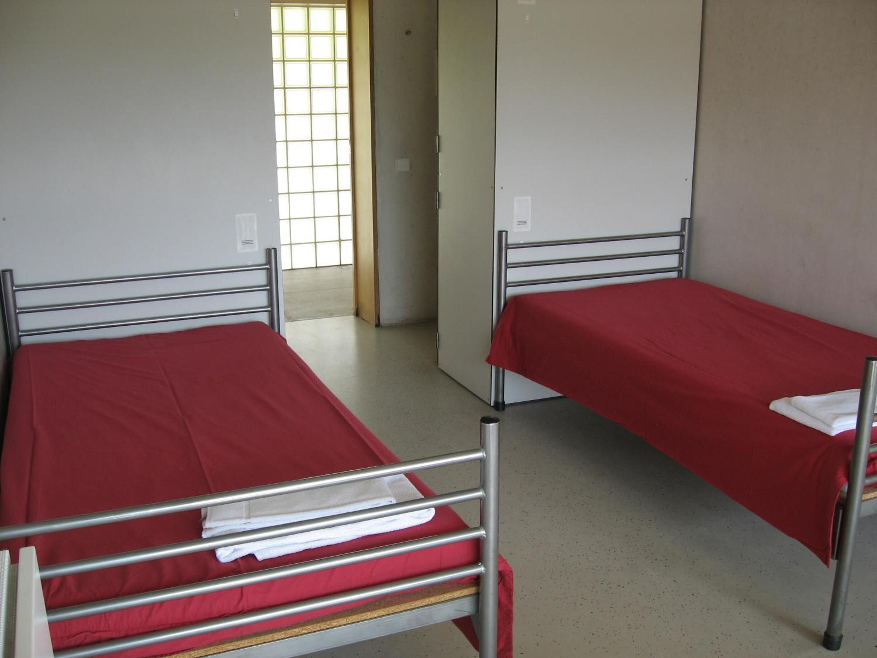 Two single beds with metal frames and red bedding in a room with white walls and a window with a grid pattern.