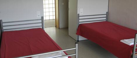 Two single beds with metal frames and red bedding in a room with white walls and a window with a grid pattern.