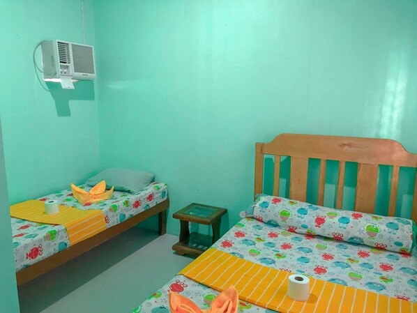 A small, brightly colored room with two beds, a small table, and an air conditioning unit on the wall. - Ging Ging Homes (Panglao)