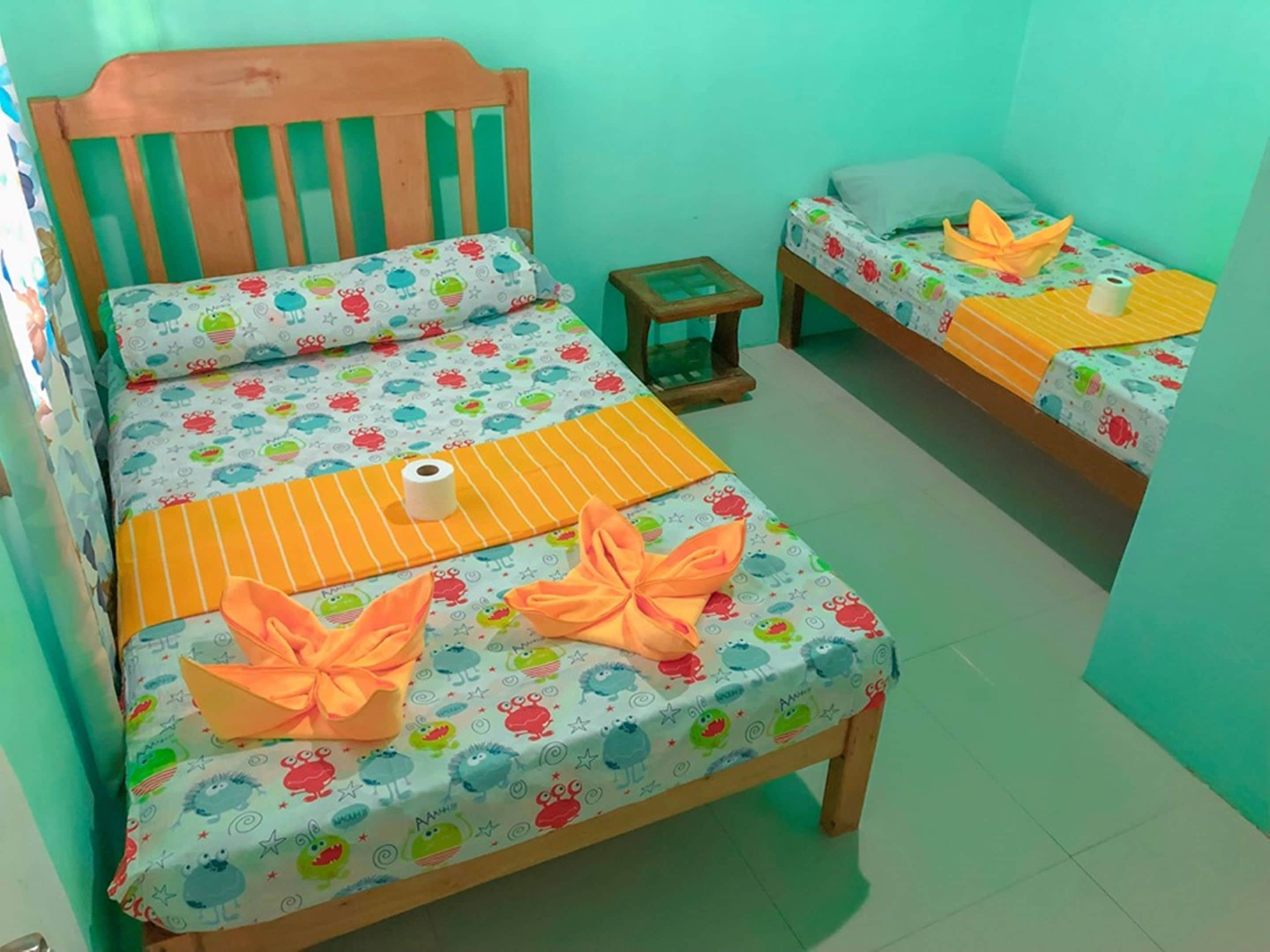 A room with two beds, each with a colorful bedspread featuring cartoon characters. There is a wooden headboard on the left bed, and a small wooden stool between the beds.