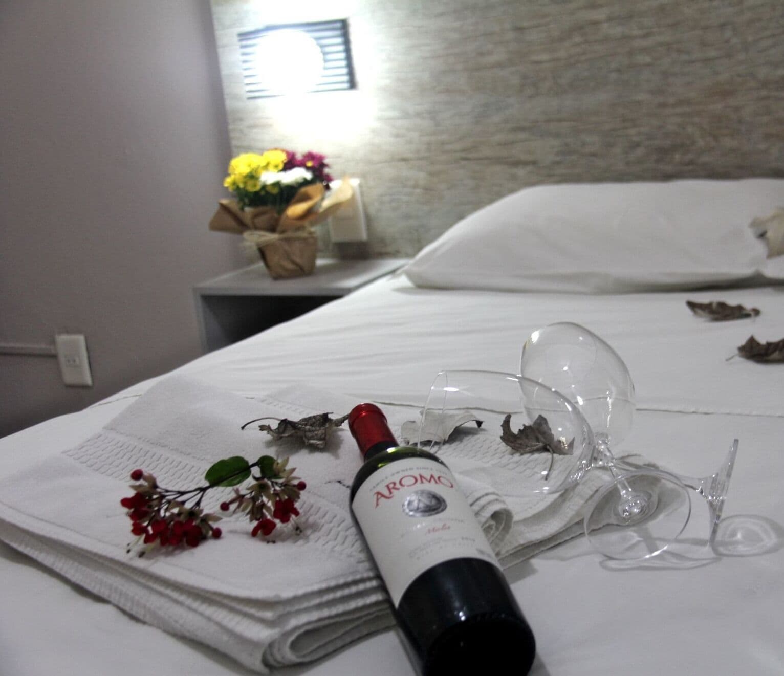 Un lit bien fait, avec une bouteille de parfum AROMA, un verre et un bouquet de fleurs posés sur un drap blanc.