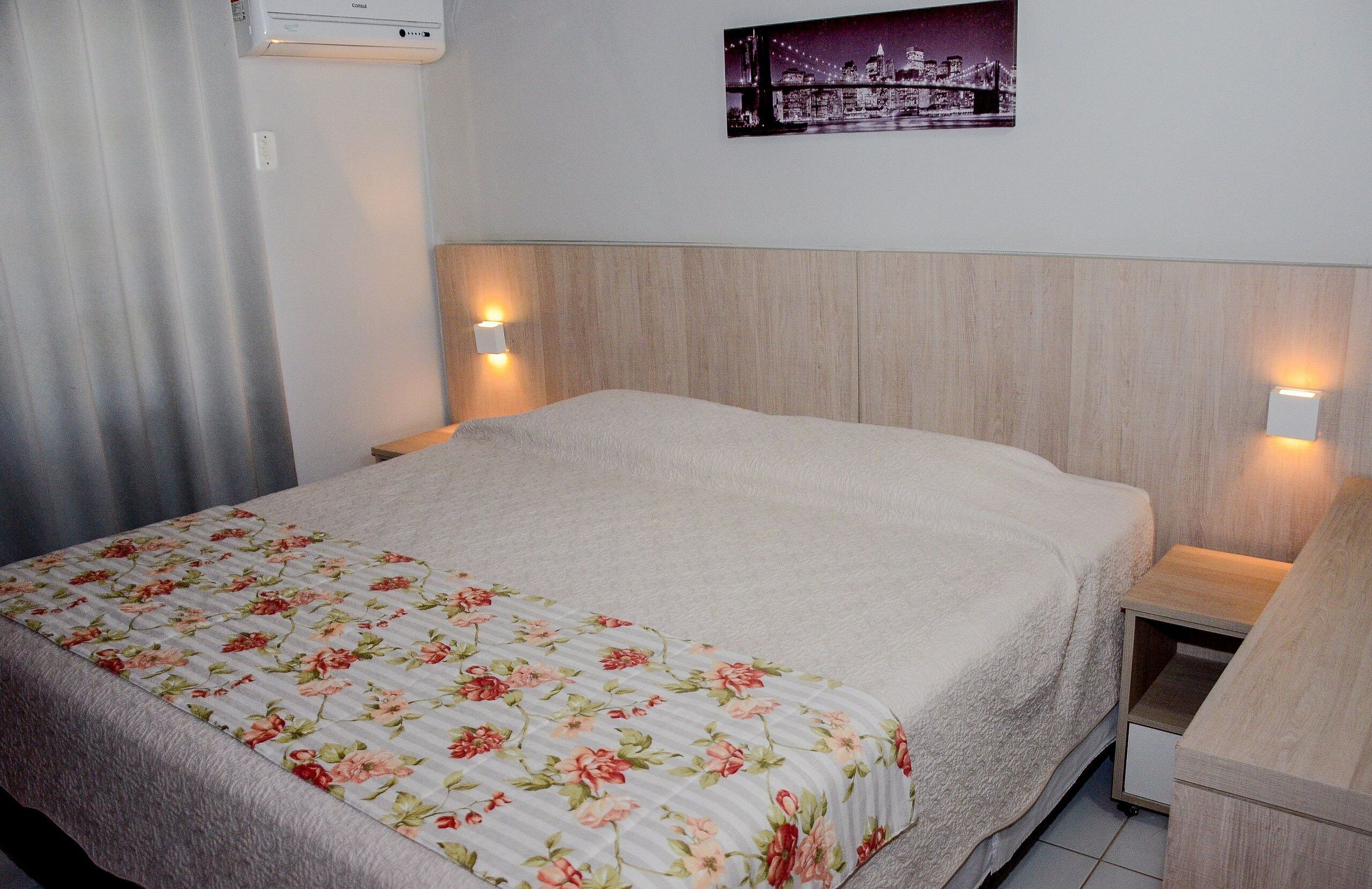 A hotel room with a bed, a nightstand, a wall-mounted air conditioner, and a cityscape picture on the wall.