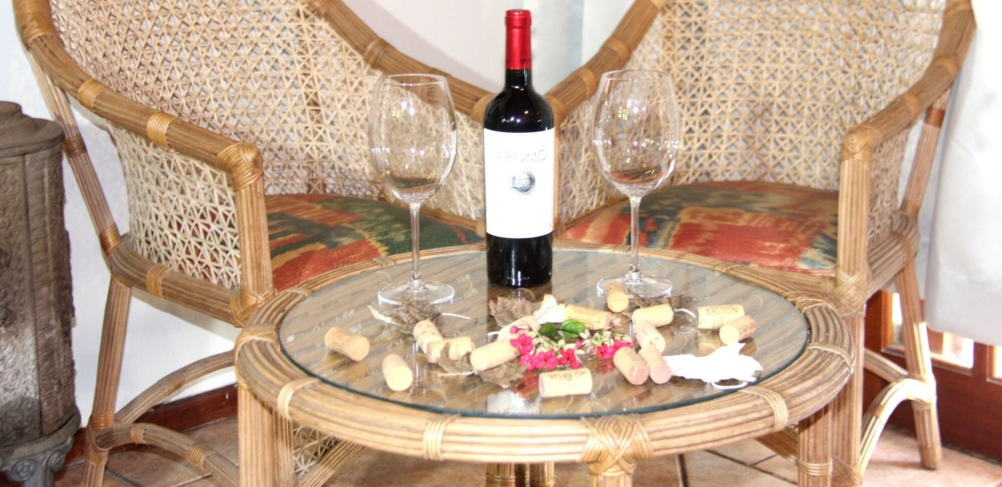 Una mesa redonda de vidrio con una botella de vino, dos copas y corchos sobre una silla de mimbre.