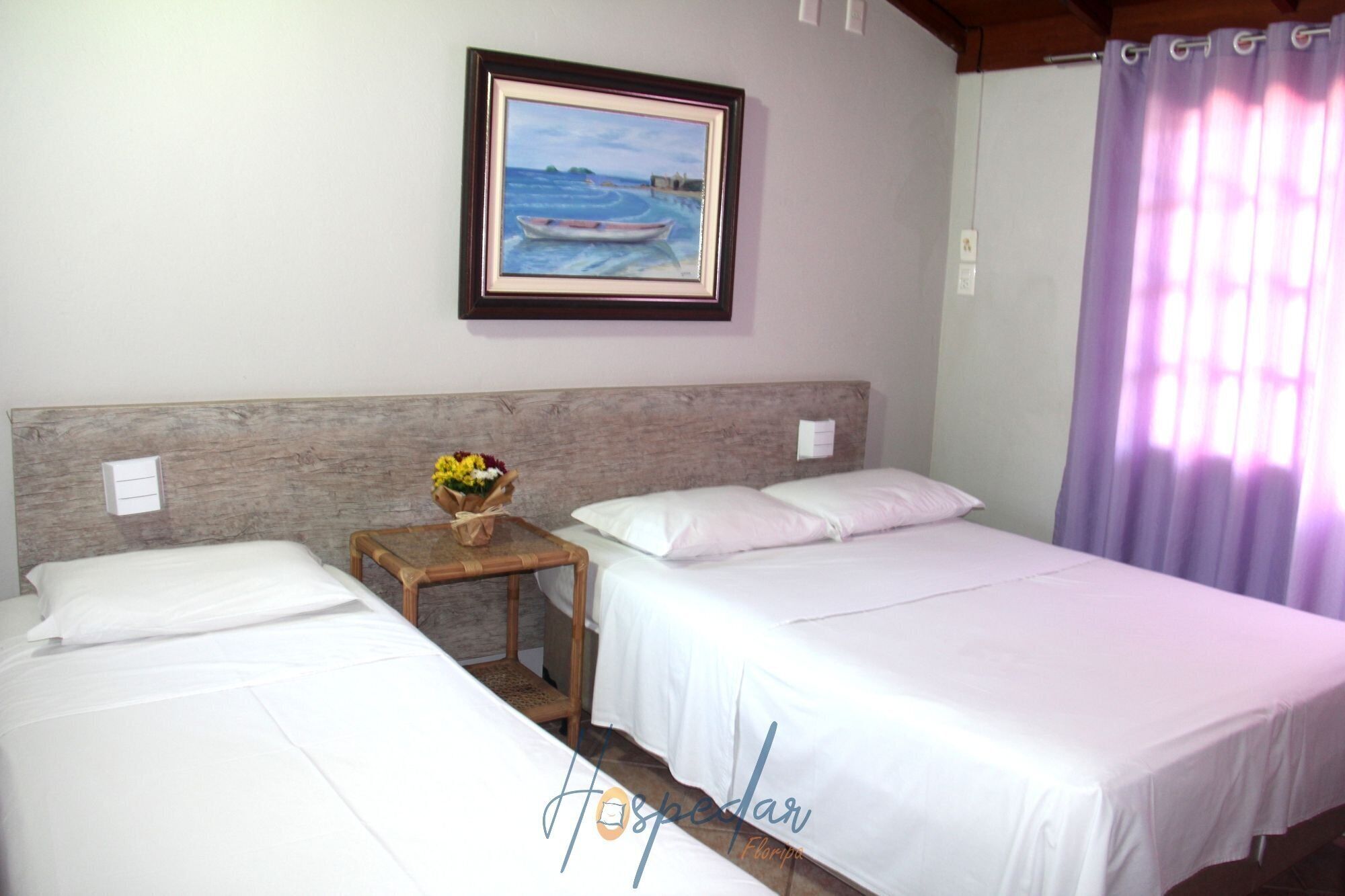 A hotel room with two beds, a wooden headboard, a framed painting of a boat, a small table with a vase of flowers, and a purple curtain.
