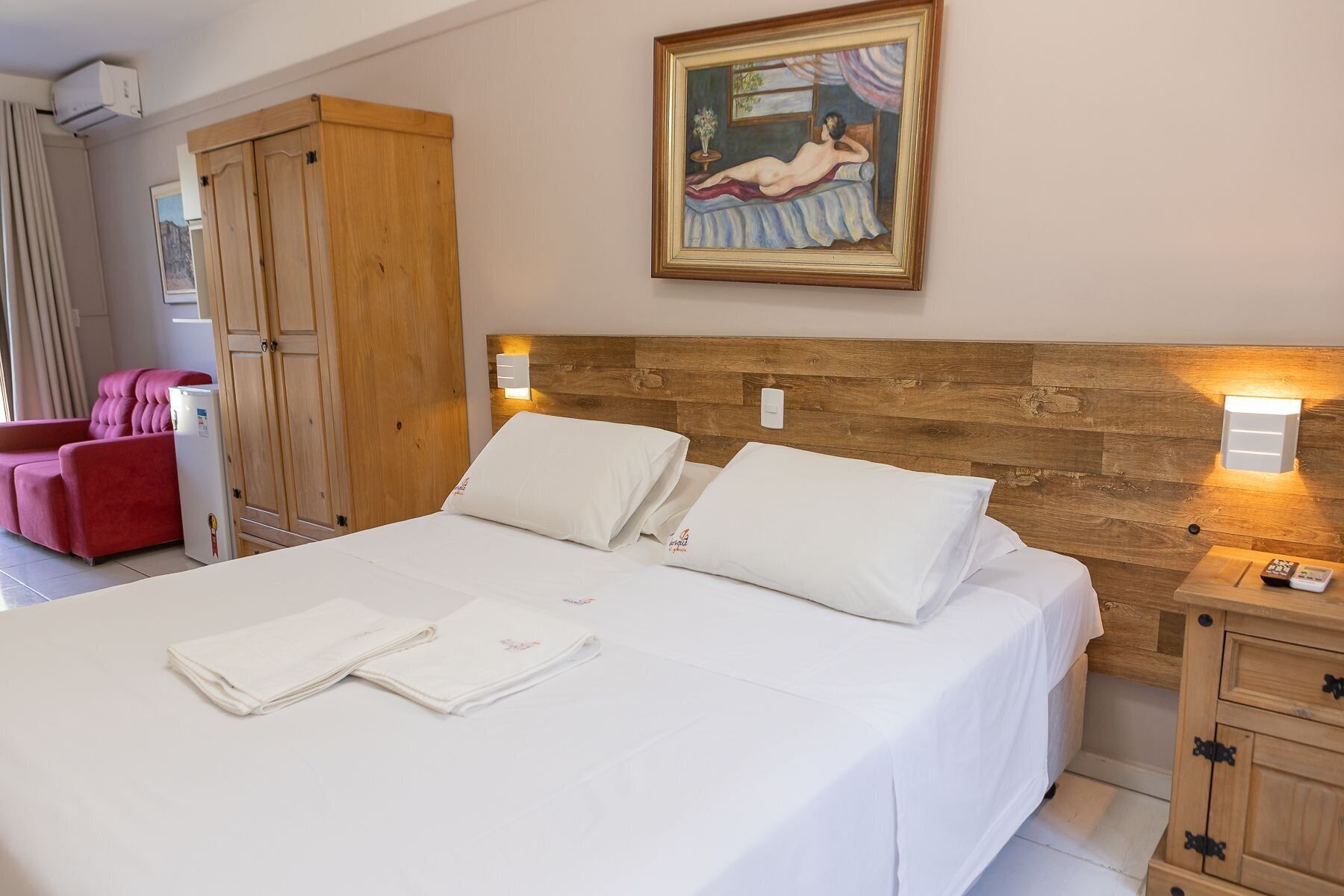 Habitació d'hotel amb un capçal de llit de fusta, un quadre damunt del llit, una cadira vermella i un armari de fusta.