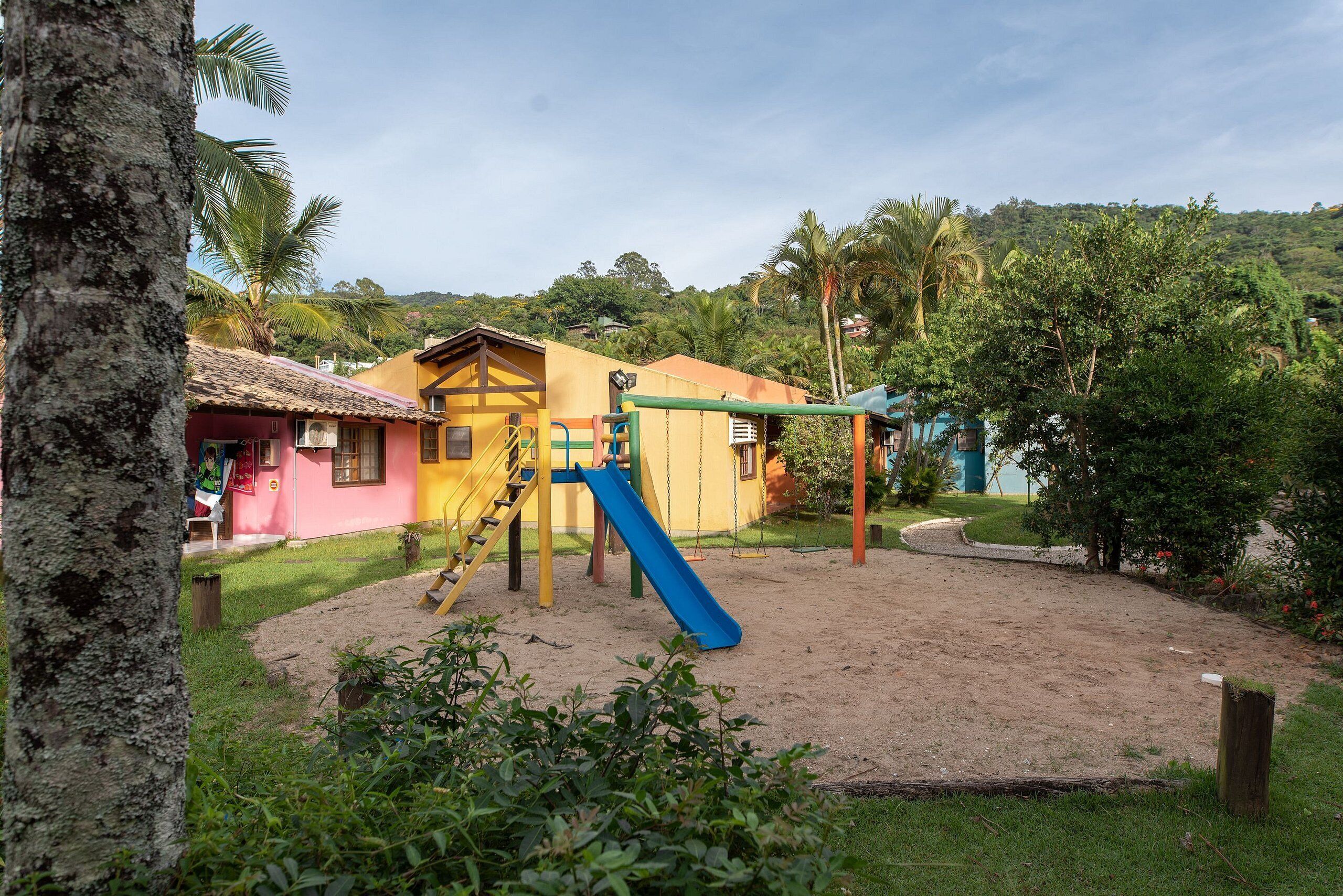 A playground with a slide and swings, surrounded by a sandy area and greenery.