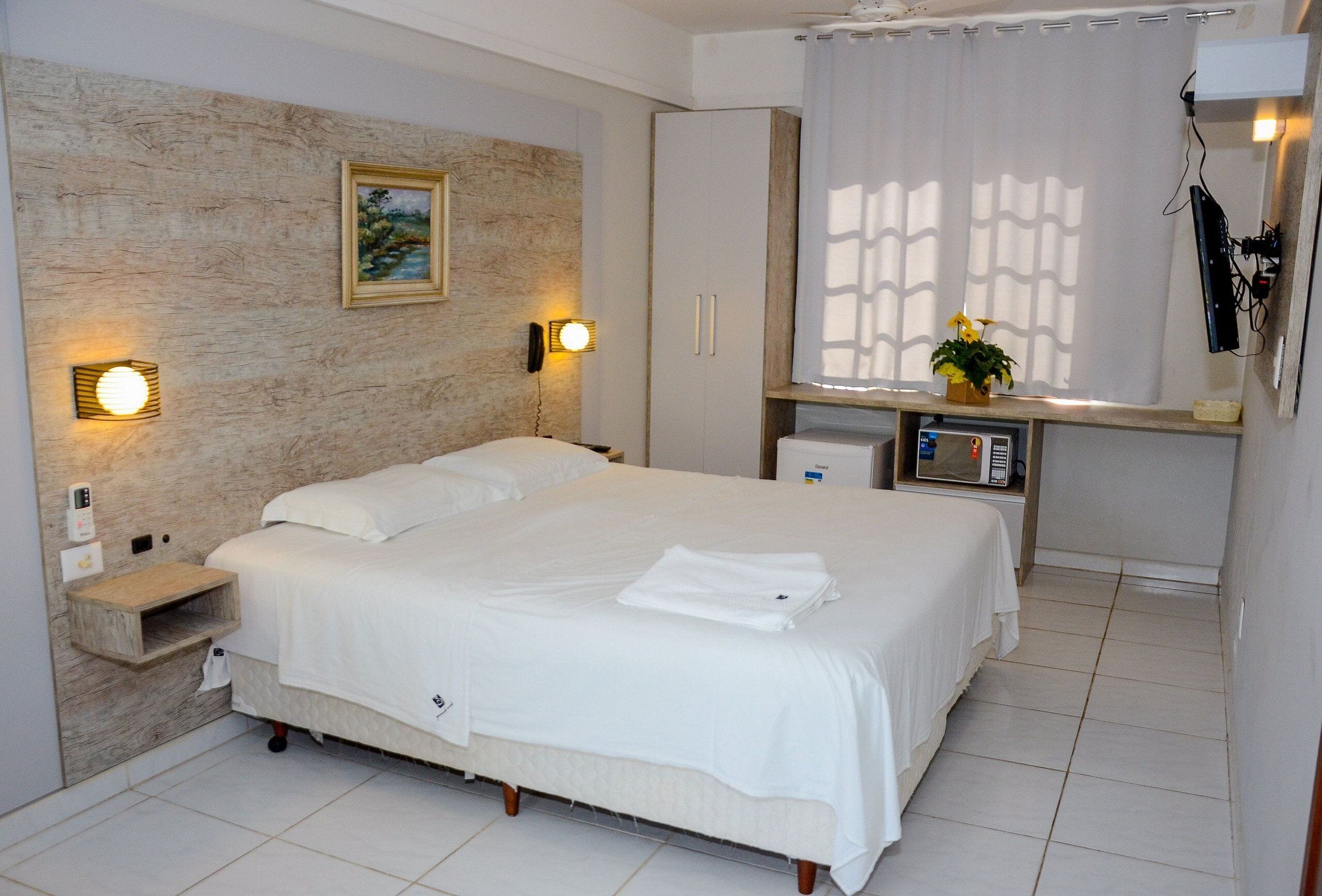 A hotel room with a large bed, bedside lamps, a painting on the wall, a small desk with a microwave, and a window with curtains.