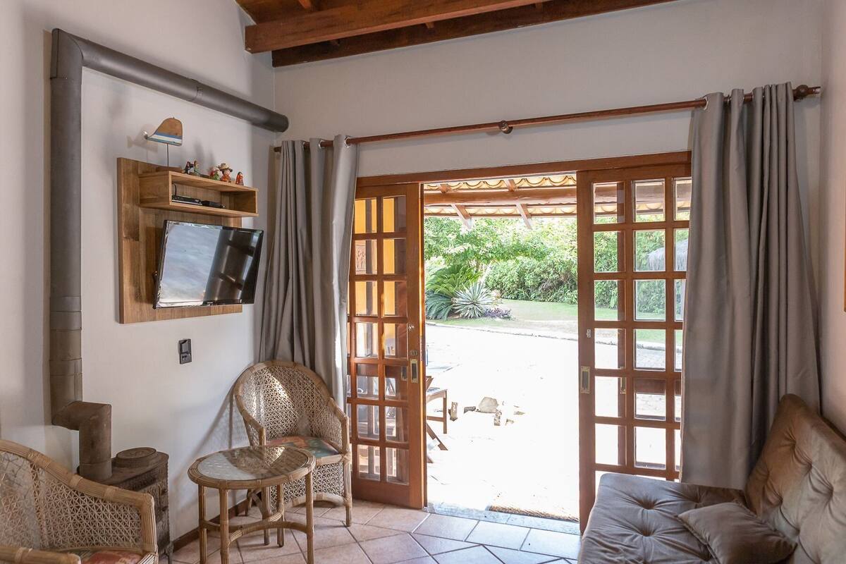 Una habitación con una televisión sobre un estante de madera, una silla de mimbre, un sofá con cojines y una gran puerta de madera que da al exterior.