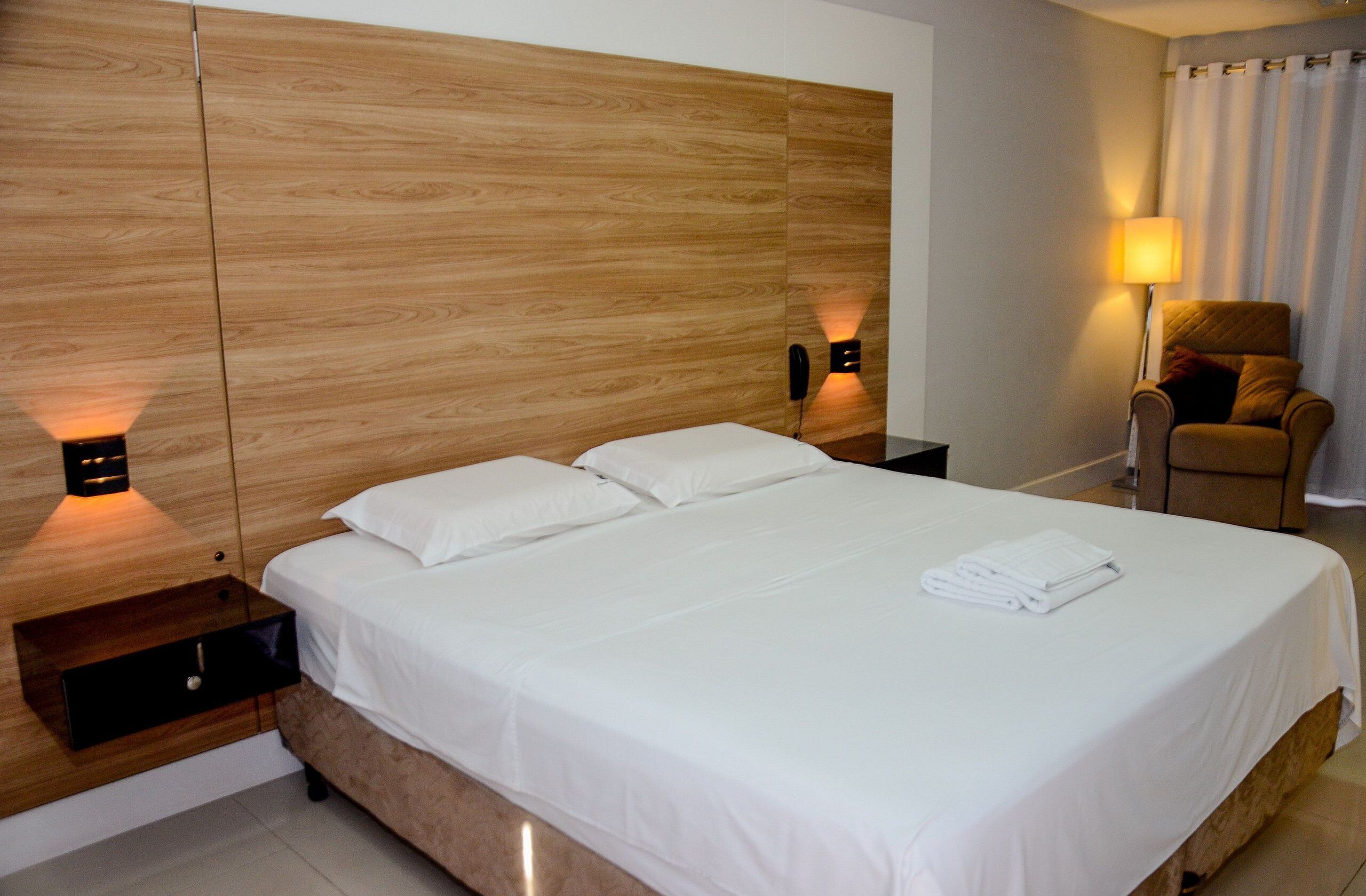 A hotel room with a wooden headboard, a bed with white linens, a nightstand with a lamp, and a chair with a cushion.