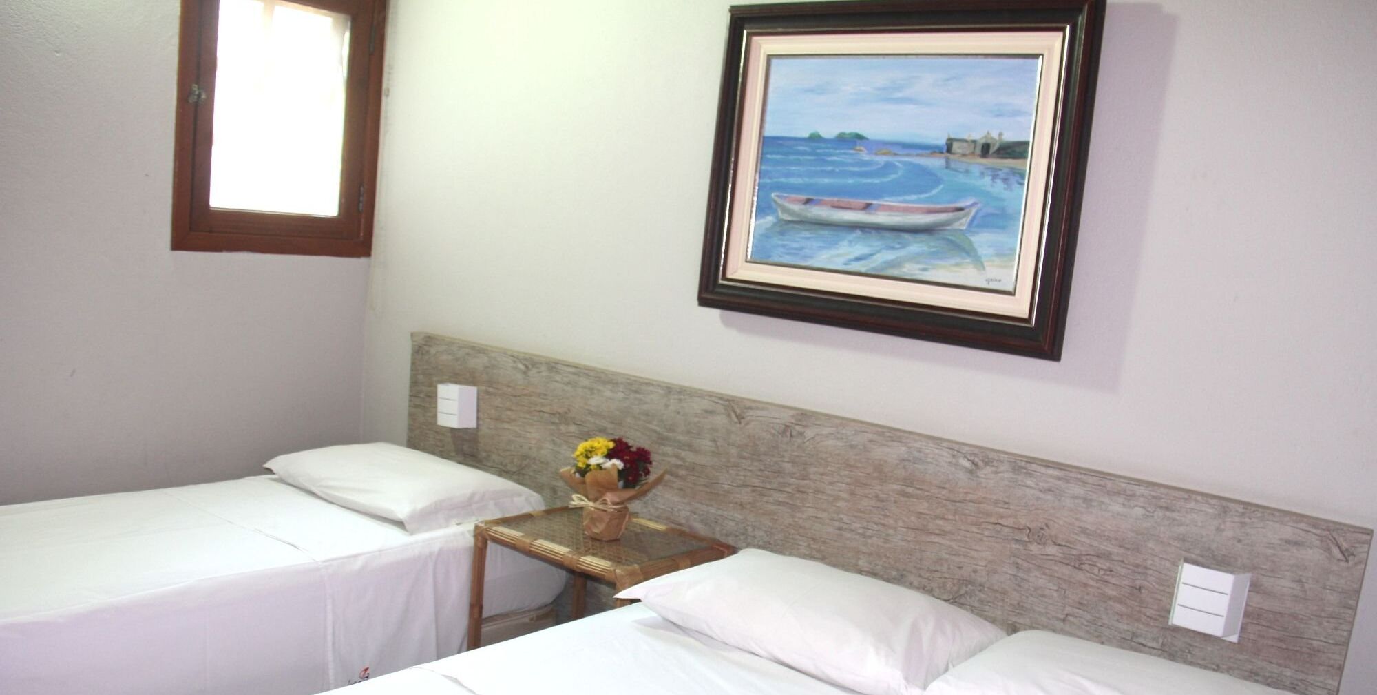 Hotelkamer met twee enkele bedden, een klein houten tafeltje met een bloevas en een schilderij van een boot aan de muur.