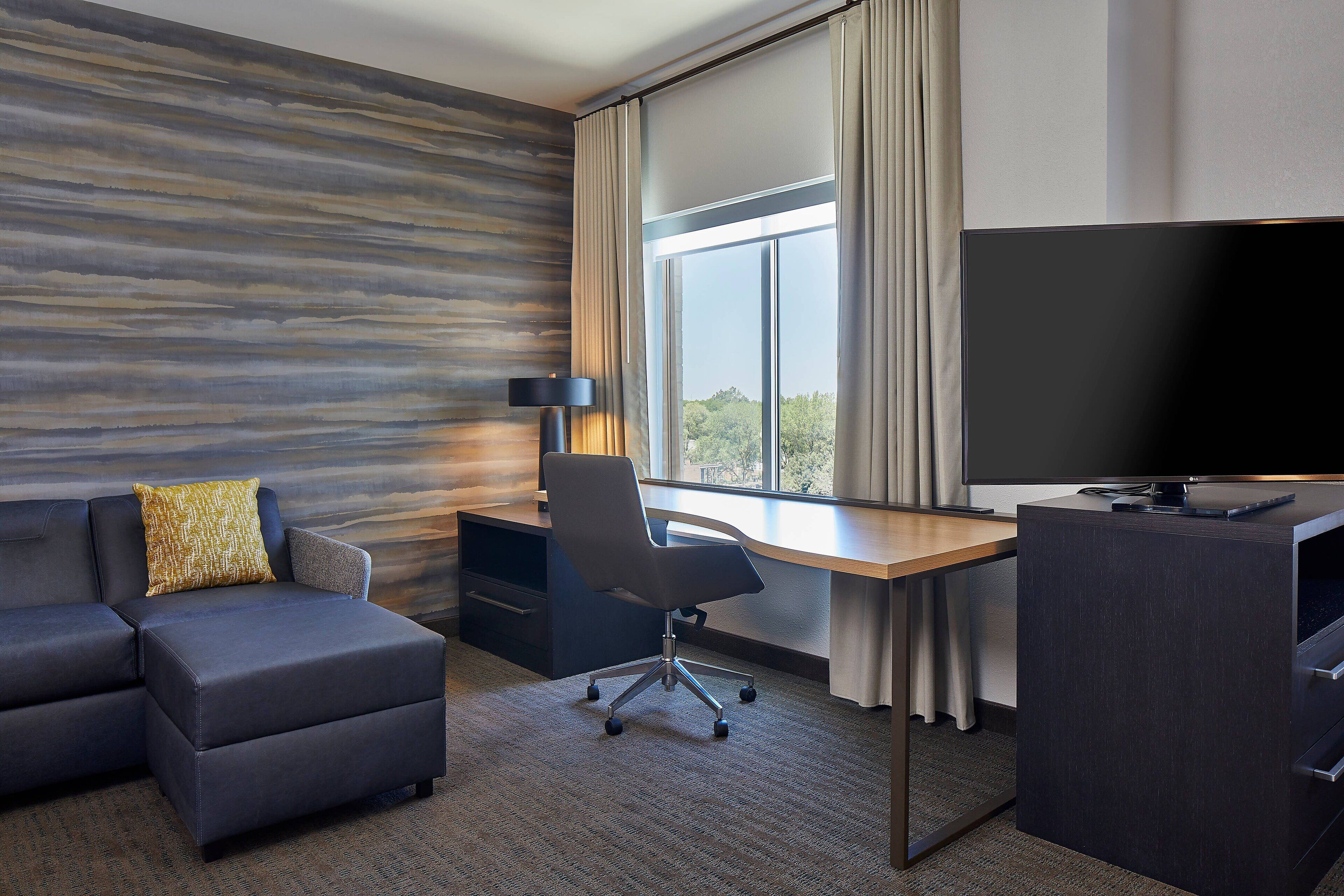 A modern hotel room with a sectional sofa, a desk with a computer, and a large flat-screen TV.