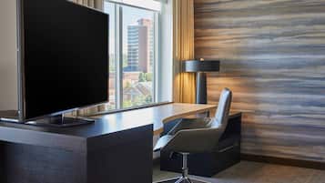 A hotel room with a large flat-screen TV on a wooden desk, a modern chair, a lamp, and a view of buildings through the window.